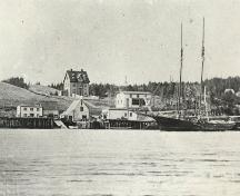 Historic photo of Reddy Premises, circa 1902.  Photo shows the Reddy Premises, including the fisheries buildings, schooners and the Reddy House prominently situated on a high hill above the water. ; HFNL 2006.