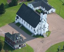 Vue aérienne de l'église Notre-Dame de l'Annonciation avec le presbytère à l'avant-plan et le Club d'âge d'or vers l'arrière.; La Société historique de la Valleé de Memramcook et le Village de Memramcook