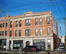 This image illustrates the front, south-facing, primary brick-clad facade of the building showing the stores at ground level with two floors of residential uses above. Note the symmetrically-designed, classically-influenced facade details.; City of Edmonton, 2004