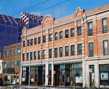 This image illustrates the front, south-facing, primary brick-clad facade of the building showing the stores at ground level with 2 floors of residential uses above. Also illustrated are the painted signs on the upper parapet.; City of Edmonton, 2004