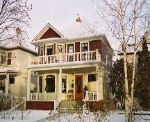 This image illustrates the two storey south and east wood-clad facades with  hipped-roof and front gable over the two-storey bay window, central chimney, full-width main floor veranda and open balcony above. (March 2004); City of Edmonton, 2004