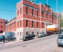 Corner view of the Armoury, showing the horizontal bands of sleek stone work, the split-faced stone foundation wall and the bartizan above the entrance which indicate the military function of the building, 1994.; Parks Canada Agency / Agence Parcs Canada, 1994.