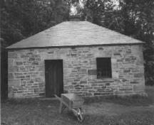 Vue de l'entrée principale de l’atelier du forgeron, qui montre sa volumétrie simple et compacte, sur un niveau et le toit en croupe à bardage en tôle, 1989.; Public Works and Government Services Canada / Travaux publics et Services gouvernementaux Canada, 1989.