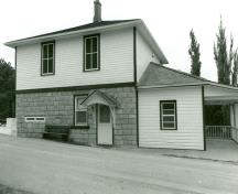 Vue de côté de la résidence fortifiée du maître-éclusier, qui montre les murs du rez-de-chaussée en maçonnerie de pierres calcaires posées en assises régulières avec des meurtrières murées et ceux de l’étage revêtus d’un parement de planches à clins, 1989; Department of Public Works / Ministère des Travaux publics, 1989.