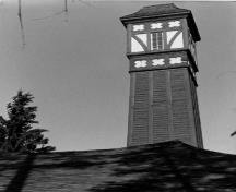 View of the Firehall's very high and prominent tower, showing the tapered tower's split logs construction, and a half-timbered cupola-like structure with bellcast pyramidal roof, 1984.; Parks Canada Agency / Agence Parcs Canada, 1984.