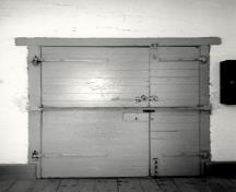 Vue de l'intérieure de la caserne, qui montre la porte en métal et la quincaillerie de la porte, 1990.; Parks Canada Agency / Agence Parcs Canada, 1990.