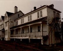 Vue en angle du bâtiment 14, qui montre la façade principale simple agrémentée de pignons à faux colombages, d’un porche à l’avant, de longues vérandas et d’escaliers de secours extérieurs.; Agence Parcs Canada / Parks Canada Agency.