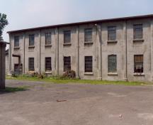 Vue de la façade de l'entrepôt et bureau, qui montre les baies des murs longs, qui sont percées par une fenêtre au rez-de-chaussée et à l’étage, 1990.; Parks Canada Agency / Agence Parcs Canada, 1990.