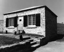 Vue de la façade générale du bâtiment 16, qui montre la volumétrie rectangulaire de plain-pied, de petite taille, coiffée d’un toit en croupe bas, 1980.; Parks Canada Agency / Agence Parcs Canada, 1980.