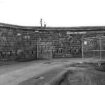 Vue générale du bâtiment 26, montrant sa façade visible incurvée revêtue de pierres taillées posées à assises irrégulières et percée de nombreuses meurtrières dans la partie inférieure du mur, qui se confond avec le mur du fossé, 1991.; Agence Parcs Canada / Parks Canada Agency, Rhona Goodspeed, 1991.