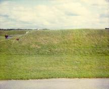 View of the side of RMC Building 30b, showing the entrance to the left and the unaltered combination of unobtrusive earthwork and minimal architectural visibility in accordance with its original purpose to store dangerous explosives, 1993.; Parks Canada Agency / Agence Parcs Canada, 1993.