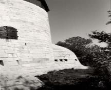 Vue de la façade latérale de la tour Cathcart, qui montre les murs extérieurs massifs, lisses et escarpés construits en blocs de calcaire serrés et deux des quatre caponnières régulièrement espacées autour de sa base, 1992.; Parks Canada Agency / Agence Parcs Canada, 1992.