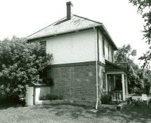 Vue en angle de la résidence fortifiée du maître-éclusier, qui montre les murs extérieurs du rez-de-chaussée réalisés en maçonnerie de pierre calcaire et ceux de l’étage revêtus d’un parement en planches à clin, 1989.; Parks Canada Agency / Agence Parcs Canada, Couture, 1989.