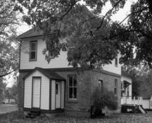Vue en angle de la résidence fortifiée du maître-éclusier, qui montre les murs extérieurs en maçonnerie de pierres posées en assises régulières du rez-de-chaussée et le parement en planches à clin de l’étage, 1990.; Parks Canada Agency / Agence Parcs Canada, James De Jonge, 1990.