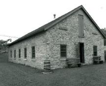 Vue en angle de la fonderie, qui montre les murs extérieurs en moellons, le positionnement de la porte et des fenêtres simplement détaillées au moyen de voussoirs et d’appuis en pierre de taille, 1989.; Public Works and Government Services Canada / Travaux publics et Services gouvernementaux Canada, 1989.