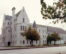 Vue en angle du manège militaire de Seaforth, où l'on peut apercevoir les toits, les deux tours circulaires avec toit conique, les contreforts massifs à décrochements, et la grosse cheminée à ressauts.; Department of National Defence / Ministère de la Défense nationale