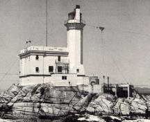 General view of the exterior of the Light Tower, showing its form and massing which consist of a tall, slightly tapered octagonal tower with narrow slit openings, flared lantern platform, lantern and light.; Parks Canada Agency / Agence Parcs Canada