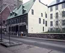 Vue de coin de la maison Maillou démontrant les murs extérieurs en maçonnerie, la pierre de taille des encadrements de portes et de fenêtres, le bandeau entre les deux étages et le revêtement de planches à clin du pignon nord-est, 1985.; Parks Canada Agency / Agence Parcs Canada, A. Guindon, 1985.