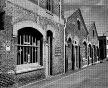 Vue des détails du bâtiment D51 et sa configuration et le traitement des portes et fenêtres avec les appuis en pierre, les arcs, les encadrements à rejetau et la haute fenestration à carreaux multiples sur tous les éléments du bâtiment, 1989.; Parks Canada Agency / Agence Parcs Canada, I. Doull, 1989.