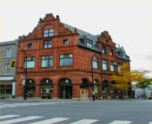 Vue de coin de la Banque de Montréal démontrant sa fenestration tel que l'arcade vitrée au rez-de-chaussée; les grandes fenêtres à l'étage; et l'éventail de fenêtres ovales, de fenêtres à guillotine et de lucarnes aux deux étages mansardés, 2004.; Parks Canada Agency / Agence Parcs Canada, 2004.