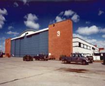 General view of Hangar 3 showing  the massive rectangular towers that house the telescoping, sliding steel doors, the two-storey flat-roofed office wings that extend along both sides of the hangar, 1999.; Department of National Defence / Ministère de la Défense nationale, 1999.