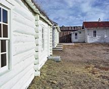 Side view of the Commissioners Residence (right) with other similar buildings (left) that demonstrate the concrete foundation and the white painted exterior walls built of regular diameter peeled logs laid horizontally, 1972.; Parks Canada Agency / Agence Parcs Canada, 1972.