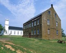 Vue générale de la Banque des fermiers de Rustico qui montre l’emplacement de l’édifice au milieu d’un ensemble d’édifices, proches de l’église St. Augustine.; Parks Canada Agency / Agence Parcs Canada.