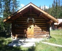 Raven Cabin Bunkhouse, front facade, showing its simple massing as a single-room with a gable roof, extended on log purlins over the entrance to shelter the stoop, 2017.; Agence Parcs Canada / Parks Canada Agency, 2017.