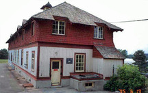 Vue en angle de l'élévation est du Bâtiment de service montrant  le parement de planches et tasseaux du rez-de-chaussée, le revêtement en bardeaux de bois de l’étage qui est légèrement évasé vers l’extérieur, 1995.; Parks Canada Agency / Agence Parcs Canada, C. Zacconi, 1995.